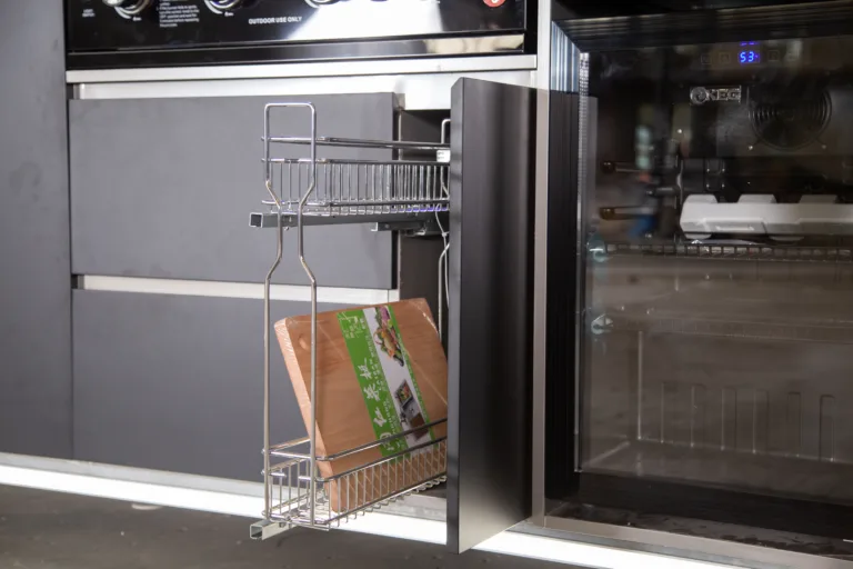 Outdoor kitchen pull-out wire basket beside a glass-door refrigerator and grill cabinet