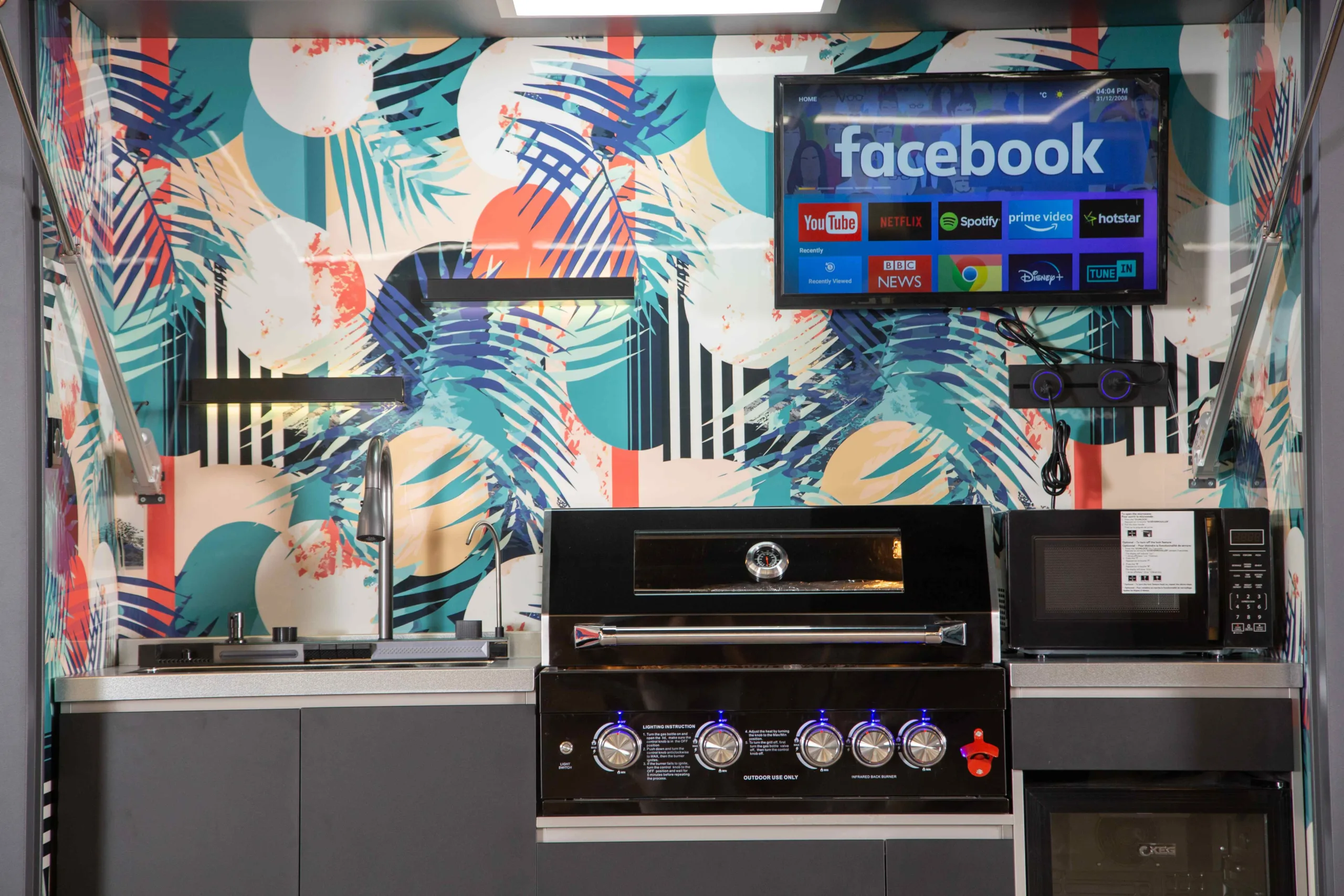 Outdoor kitchen with colorful tropical-pattern backsplash, grill, sink, refrigerator, television, and lighting